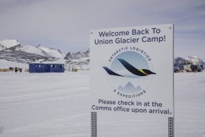 A sign reading Welcome Back To Union Glacier Camp! Antarctic Logistics and Expeditions. Please check in at the Comms office upon arrival stands in snowy Antarctica, with tents and mountains in the background near the South Pole.