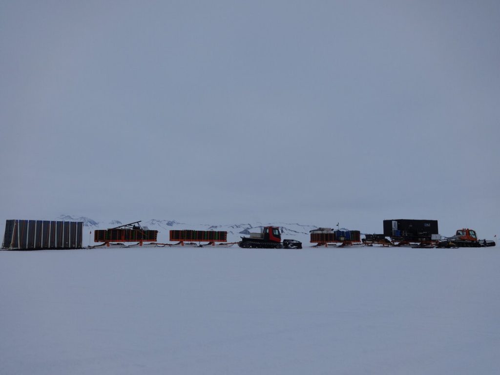 Several tracked vehicles haul large sledges carrying equipment and containers across a vast, snowy landscape in Antarctica, with distant mountains visible under an overcast sky—a scene typical of Antarctic Logistics and Expeditions near the South Pole.