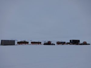 Several tracked vehicles haul large sledges carrying equipment and containers across a vast, snowy landscape in Antarctica, with distant mountains visible under an overcast sky—a scene typical of Antarctic Logistics and Expeditions near the South Pole.