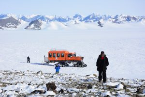Three people stand on a snowy, rocky landscape in Antarctica with an orange tracked vehicle nearby; snow-capped mountains rise in the background under a clear sky.