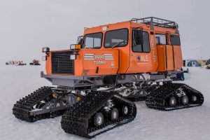 An orange, tracked snow vehicle with four sets of treads is parked on snowy ground in Antarctica. Equipped with windows, headlights, and a roof rack, it’s built for Antarctic Logistics and Expeditions in harsh icy conditions.