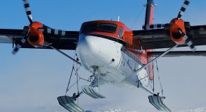 A red and white twin-propeller airplane with skis attached to its landing gear, operated by Antarctic Logistics and Expeditions, flies low over a snowy Antarctica landscape, its propellers spinning and the sky clear in the background.