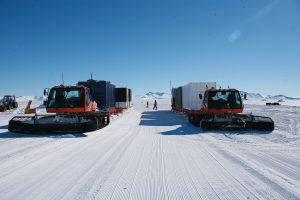 Two large orange snow vehicles from Antarctic Logistics and Expeditions tow containers across a snowy, flat landscape under a clear blue sky in Antarctica. Mountains rise in the distance as a person stands between the vehicles near the South Pole.