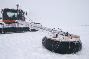 An orange snow vehicle operated by Antarctic Logistics and Expeditions pulls a metal frame attached to a rubber sled over the snowy landscape of Antarctica, likely for scientific research; distant mountains rise in the background.