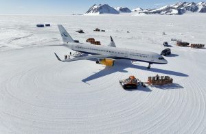 A large passenger airplane sits on a snowy, icy runway in Antarctica, surrounded by snow vehicles and mountains in the background. Operated by Antarctic Logistics and Expeditions, people and cargo are visible near the aircraft.
