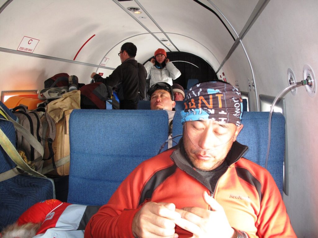 A man in an orange jacket sits near a window inside a small plane en route to the South Pole, focusing on his hands. Behind him, other Antarctic Logistics and Expeditions passengers and backpacks are visible, with two people talking at the back.