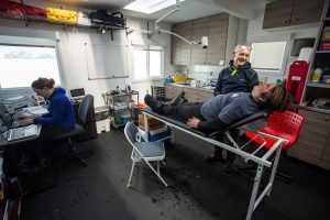 In a lab near the South Pole, a person lies on an examination table while another stands nearby, both smiling. A third person works on a laptop in the background. Cabinets, medical equipment, and supplies fill the Antarctic Logistics and Expeditions room.