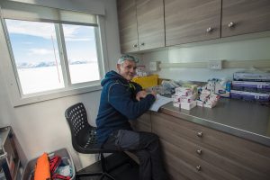 A person wearing glasses and a blue hoodie sits at a desk in a medical office with supplies, smiling at the camera. A snowy Antarctic landscape and distant mountains recall the remote beauty of Antarctica and the spirit of South Pole expeditions.