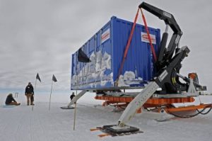 A blue shipping container with a crane arm and skis is stationed on snowy ground in Antarctica. Three people stand or sit nearby, with small black flags marking the area under an overcast sky—part of Antarctic Logistics and Expeditions.