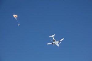 A small airplane flies in a clear blue sky above Antarctica, towing an orange and white parachute that trails behind it—a striking scene during Antarctic Logistics and Expeditions flights near the South Pole.