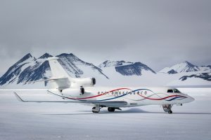 A private jet with colorful wave designs on its fuselage is parked on a snowy, icy plain in Antarctica, with snow-covered mountains in the background under an overcast sky, supported by Antarctic Logistics and Expeditions.
