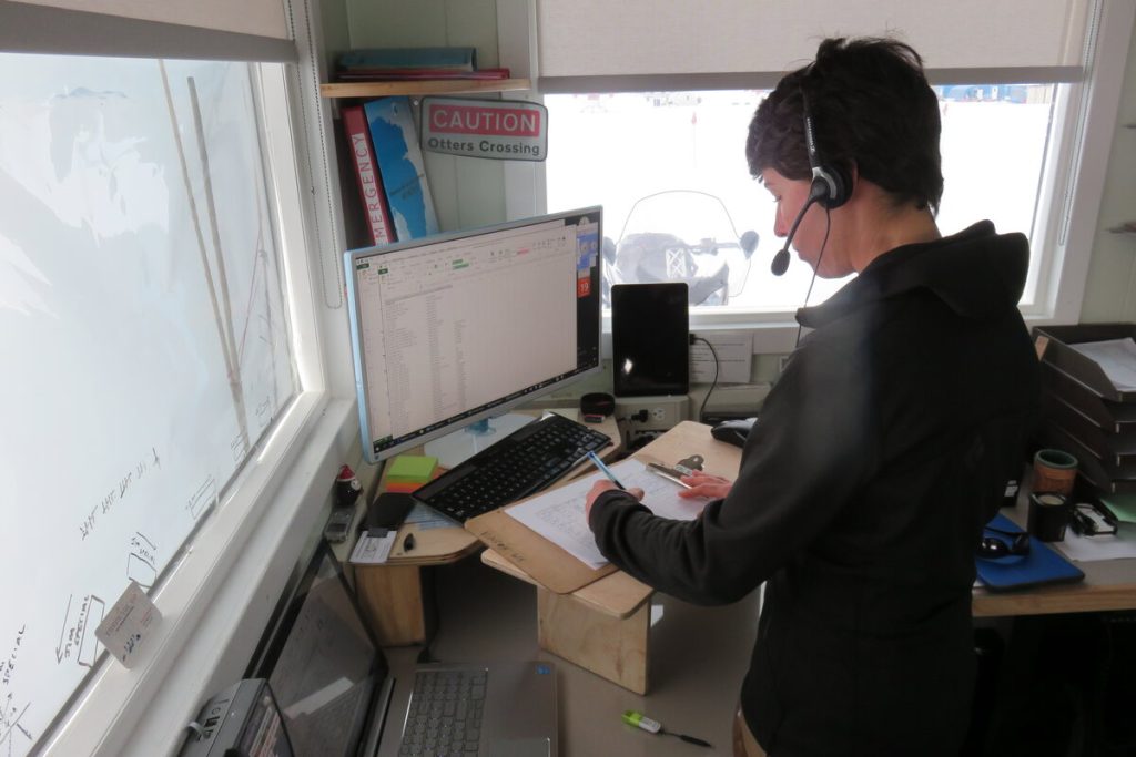 A person wearing a headset stands at a desk, looking at a computer monitor with spreadsheets—perhaps tracking Antarctic Logistics and Expeditions. They write on a clipboard, with office supplies and a “Caution Otters Crossing” sign in the background.