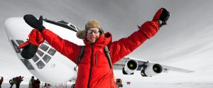 A person in a red parka, winter hat, and sunglasses smiles with arms raised in front of a large white airplane on a snowy landscape in Antarctica. Other people and Antarctic Logistics and Expeditions equipment are visible in the background.