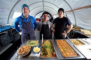 Three people wearing aprons and hats stand behind a buffet table inside a tent in Antarctica, serving hot dishes like pasta, vegetables, and baked casseroles—a snapshot of daily life with Antarctic Logistics and Expeditions at the South Pole.