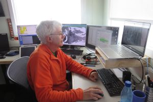 An older man in an orange jacket works at a desk with multiple computer monitors displaying maps and data from Antarctica. He uses a desktop keyboard and mouse, with a laptop propped up on a box. A water bottle sits nearby.