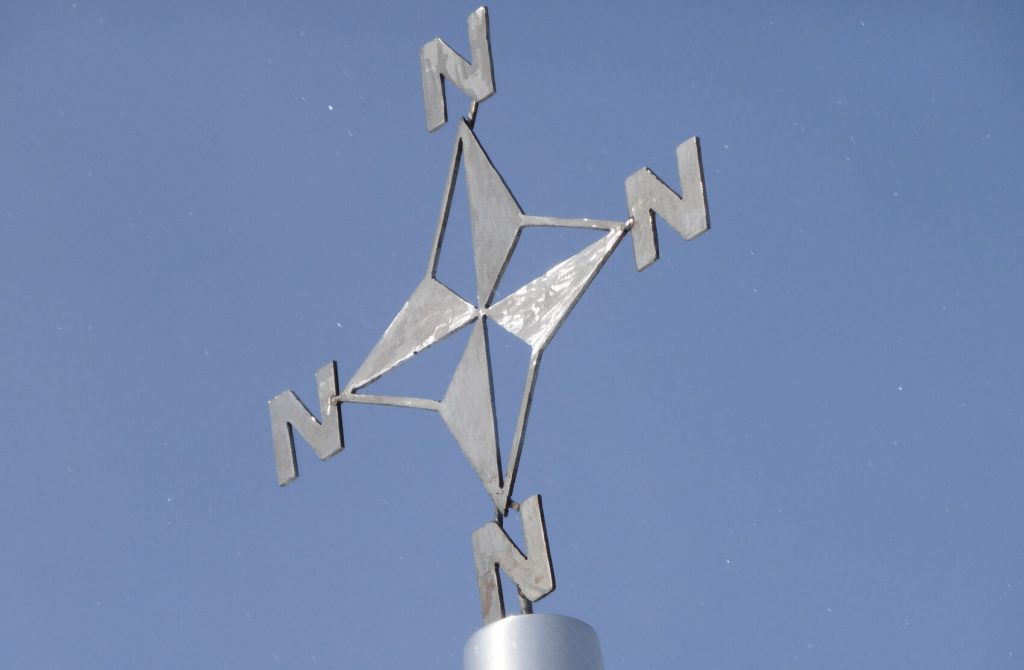 A metallic weather vane with four arrows, each labeled N, against a clear blue sky. The design suggests every direction points north—an homage to the unique geography of the South Pole in Antarctica.