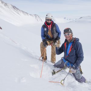 Two climbers in winter gear smile while kneeling in deep snow on a snowy mountain slope, holding ropes and ice axes—ready for their next Antarctica adventure—with snowy peaks and a cloudy sky in the background.