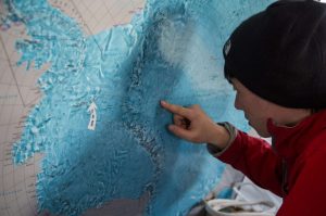 A person in a red jacket and black beanie points at a location near the South Pole on a large blue and white map, closely examining the details, possibly planning with Antarctic Logistics and Expeditions.