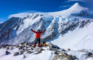 A climber in red winter gear stands triumphantly on a snowy, rocky mountain slope in Antarctica, with arms raised. Surrounded by snow-covered peaks and a bright blue sky, they celebrate reaching new heights with Antarctic Logistics and Expeditions.