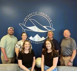 Six people smile while standing and sitting in front of a blue wall with the Antarctic Logistics & Expeditions logo, reflecting their passion for Antarctica. A globe is placed on a table in front of them.