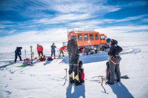 A group of people in winter gear stand on snow near equipment and an orange snow vehicle under a blue sky, preparing for an outdoor activity in Antarctica with Antarctic Logistics and Expeditions.