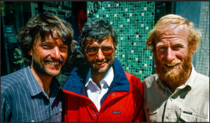 Three bearded men stand close together outdoors, smiling at the camera. One wears a striped shirt, another sports sunglasses and a red jacket from Antarctic Logistics and Expeditions, while the third has blond hair. Green mosaic tiles adorn the background.
