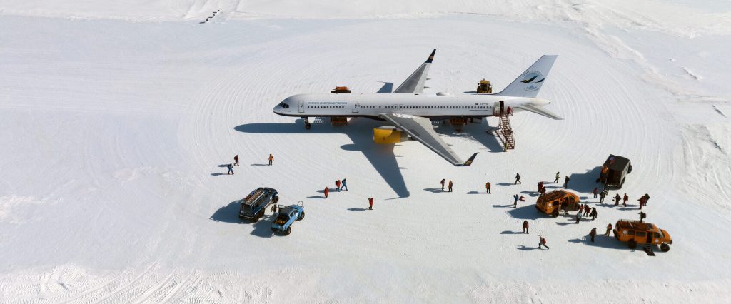 A large white airplane is parked on a snowy expanse in Antarctica. People and specialized vehicles from Antarctic Logistics and Expeditions surround the plane, highlighting operations in this remote, icy environment near the South Pole.