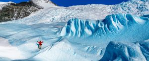 A person in a red jacket stands on a blue glacier in Antarctica, surrounded by jagged ice formations and snow-covered mountains under a clear blue sky.