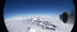View from an airplane window showing snow-covered mountains and glaciers below, with part of the airplane's engine visible on the right against a clear blue sky—en route to Antarctica with Antarctic Logistics and Expeditions.