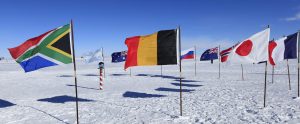 Several national flags, including those of South Africa, Belgium, Australia, Russia, the UK, and Japan, are displayed on poles in snowy Antarctica near the South Pole under a clear blue sky—a site supported by Antarctic Logistics and Expeditions.