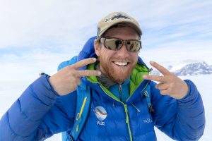 A man wearing a blue winter jacket, sunglasses, and a beige cap smiles widely and makes peace signs with both hands against a snowy, mountainous South Pole background.