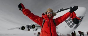 A person in a red winter jacket and fur hat smiles with arms raised at the South Pole, standing before a large airplane on Antarctica’s snowy landscape, with other people and Antarctic Logistics and Expeditions vehicles in the background.