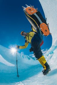 A mountaineer in bright gear scales an icy slope in Antarctica, wearing crampons and holding a pole. The photo, taken from below, captures the sun shining in a clear blue sky with ice reflecting around this Antarctic Logistics and Expeditions scene.