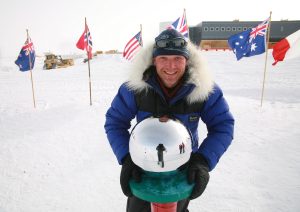A smiling person in a blue parka with a fur hood stands outdoors in snow, holding a shiny silver sphere that reflects their image and surrounding flags, with polar station buildings in the background.