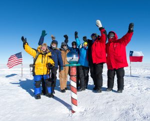 A group of people in winter gear wave at the South Pole, standing next to a striped pole and reflective sphere. Behind them are the U.S. and Chilean flags on Antarctica’s snowy landscape under a clear blue sky.