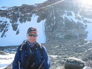 A person in outdoor gear, sunglasses, and a cap stands on rocky ground with patches of snow, in front of a tall, steep, snow-dusted mountain cliff under bright sunlight.