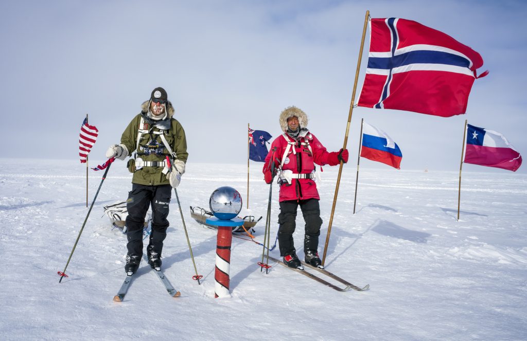 Two people in cold-weather gear stand on the snowy landscape of Antarctica at the South Pole, holding ski poles. They are surrounded by several national flags, including Norway, USA, Russia, New Zealand, and Chile.