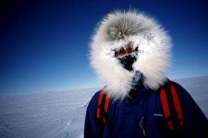A person wearing a blue parka with a fur-lined hood, red backpack straps, and icicles forming around their face stands in a snowy, frozen landscape under a clear blue sky.
