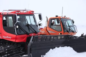 Two snowcats with drivers inside, one red and one orange, are parked side by side on the snowy landscape of Antarctica. Both vehicles, equipped with large front snowplows, await their next task under an overcast South Pole sky.