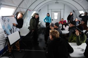 A group of people in winter clothing sit inside a white tent, listening to a person pointing at a large map on an easel at the front of the room. Some people are taking notes, while others watch attentively.