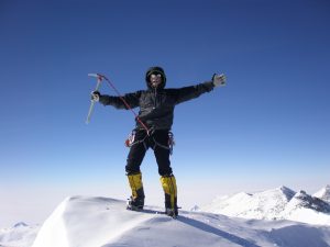 A climber wearing cold-weather gear and sunglasses stands on a snow-covered Antarctic peak with arms outstretched, holding an ice axe, surrounded by clear blue sky and the pristine mountains of Antarctica in the background.
