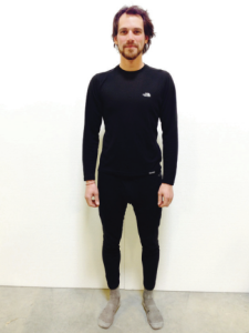 A man stands against a plain white background, wearing a black long-sleeve shirt and black pants with grey socks—dressed as if ready for an Antarctic Logistics and Expeditions journey to the South Pole. He is facing forward and smiling slightly.