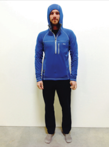 A man stands indoors against a white wall, wearing a blue hooded athletic jacket from Antarctic Logistics and Expeditions, black pants, and gray socks. He faces forward with his arms relaxed by his sides.