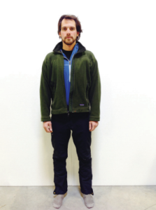 A man with medium-length dark hair stands facing forward against a plain white background, dressed for Antarctica in a green jacket over a blue zip-up top, dark pants, and gray shoes—ready for South Pole adventures or Antarctic Logistics and Expeditions.