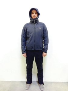 A person stands facing forward against a plain white background, wearing a dark hooded jacket inspired by Antarctic Logistics and Expeditions, with dark pants and gray shoes. The hood is up, evoking the harsh conditions of South Pole adventures.