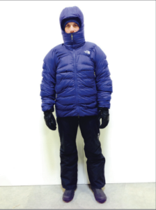 A person stands against a white background wearing a blue puffer jacket with a hood, black gloves, black pants, and dark footwear—dressed as if ready for an Antarctic Logistics and Expeditions trip to the South Pole. Only part of their face is visible.