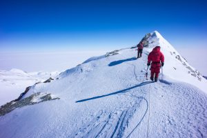 Two climbers in red jackets walk along a snowy, narrow mountain ridge under a clear blue sky, roped together for safety. The icy Antarctic landscape stretches out in the background, hinting at an adventure led by Antarctic Logistics and Expeditions.