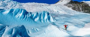 A person in a red jacket stands triumphantly with raised arms on rugged, blue ice formations of an Antarctic glacier, surrounded by snow-covered peaks under a clear blue sky.