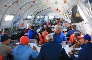 People in winter clothing eat together inside a decorated tent with hanging ornaments at the South Pole. The tent is brightly lit, and everyone appears to be enjoying a communal meal at long tables in Antarctica.