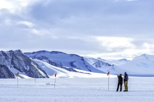 Two people stand on a snowy Antarctic landscape, surrounded by red-flag markers. Mountains and glaciers rise in the background, under a cloudy blue sky—a scene reminiscent of an expedition with Antarctic Logistics and Expeditions.